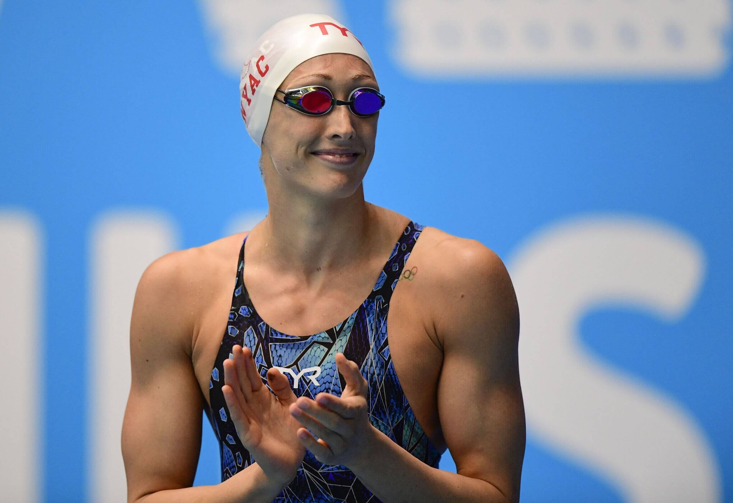Breeja Larson clapping her hands with a blue swim suit, white swimming cap and goggles